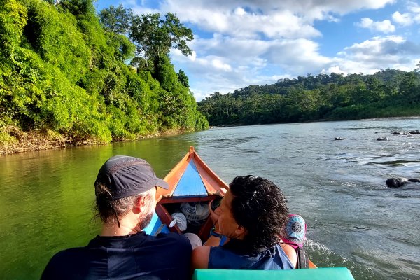 Two people riding in a small wooden boat on a calm river surrounded by lush green rainforest under a blue sky with scattered clouds.
